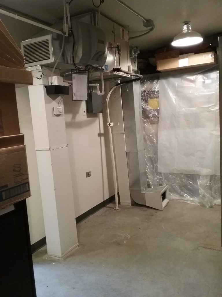 Cleared area in Collections Room 1 of The Charles A. Weyerhaeuser Memorial Museum, Little Falls, MN. The small HVAC unit that will be replaced as part of the project is shown near the top of this photo, with shelves of collections items next to it covered in plastic. Photo taken October 2, 2021.